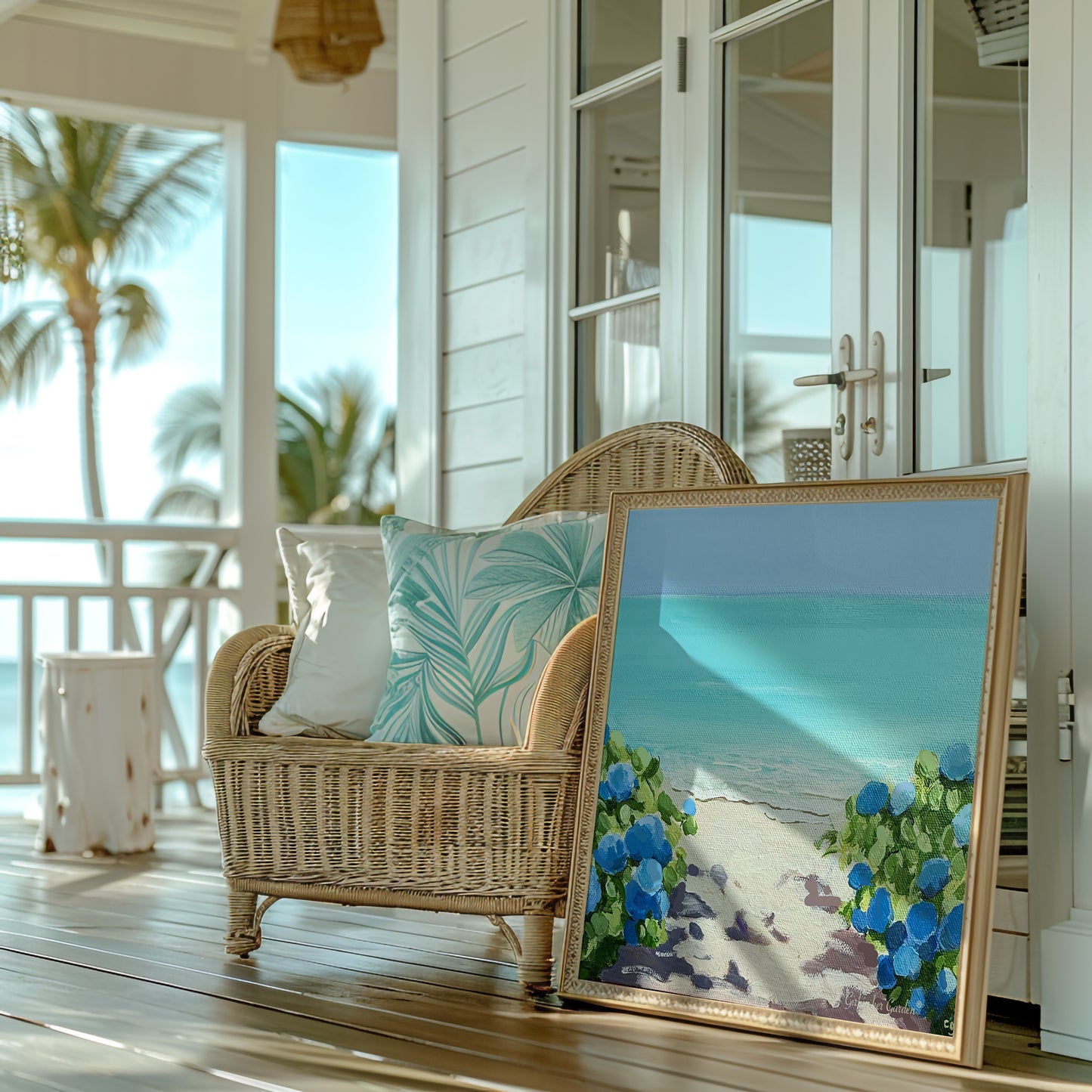A framed beach art print leaning against a wicker chair on a sunlit porch. The print depicts a turquoise ocean, white sand, and blue flowers, while the chair has white and teal patterned cushions.