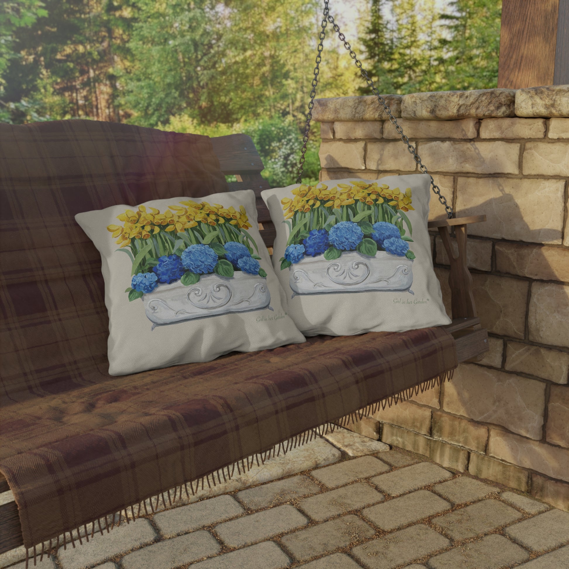 Decorative pillows with floral design on a wooden swing against a stone wall and greenery background.
