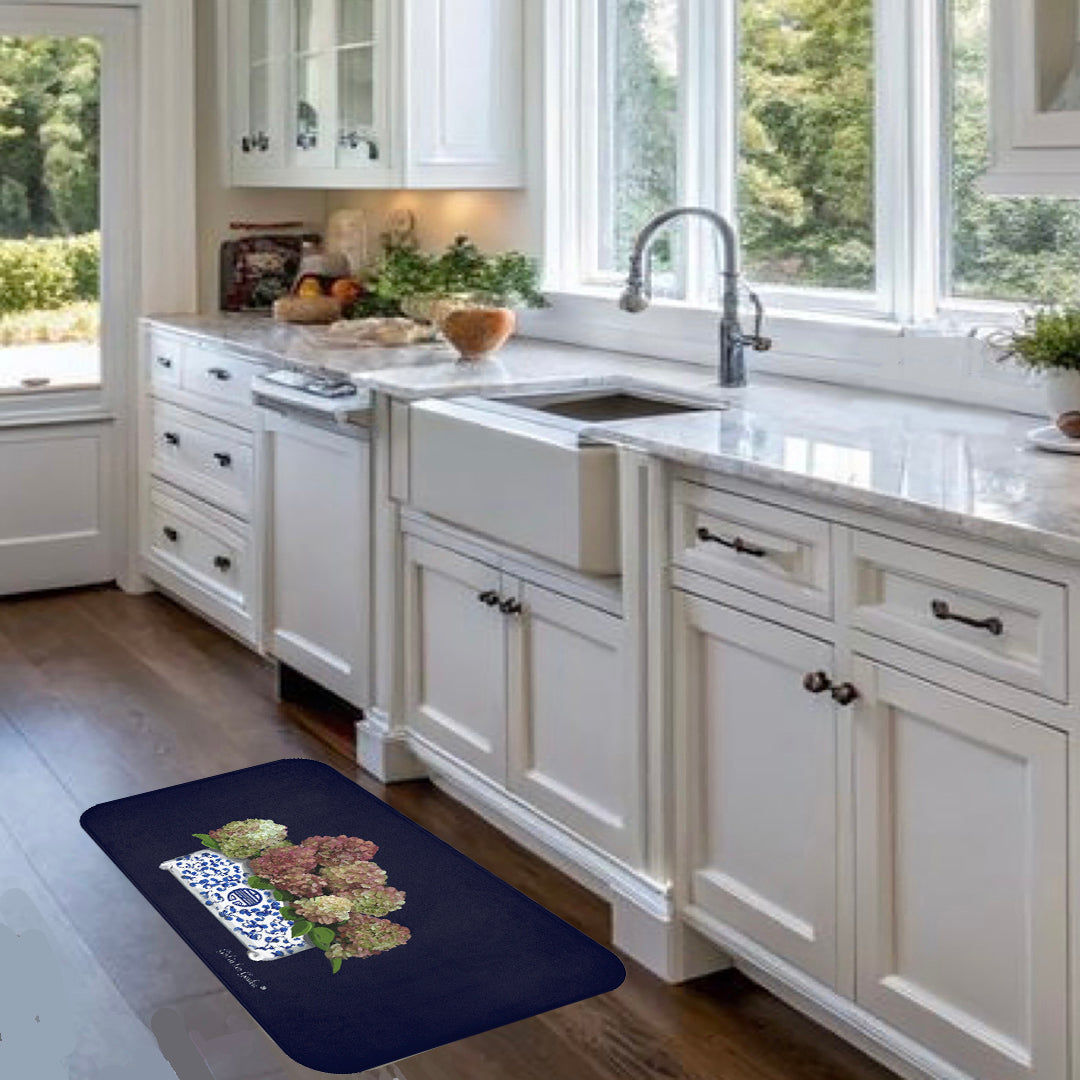 A dark navy blue rectangular kitchen mat with a floral and blue patterned design, placed on a dark wood floor in a bright white kitchen.