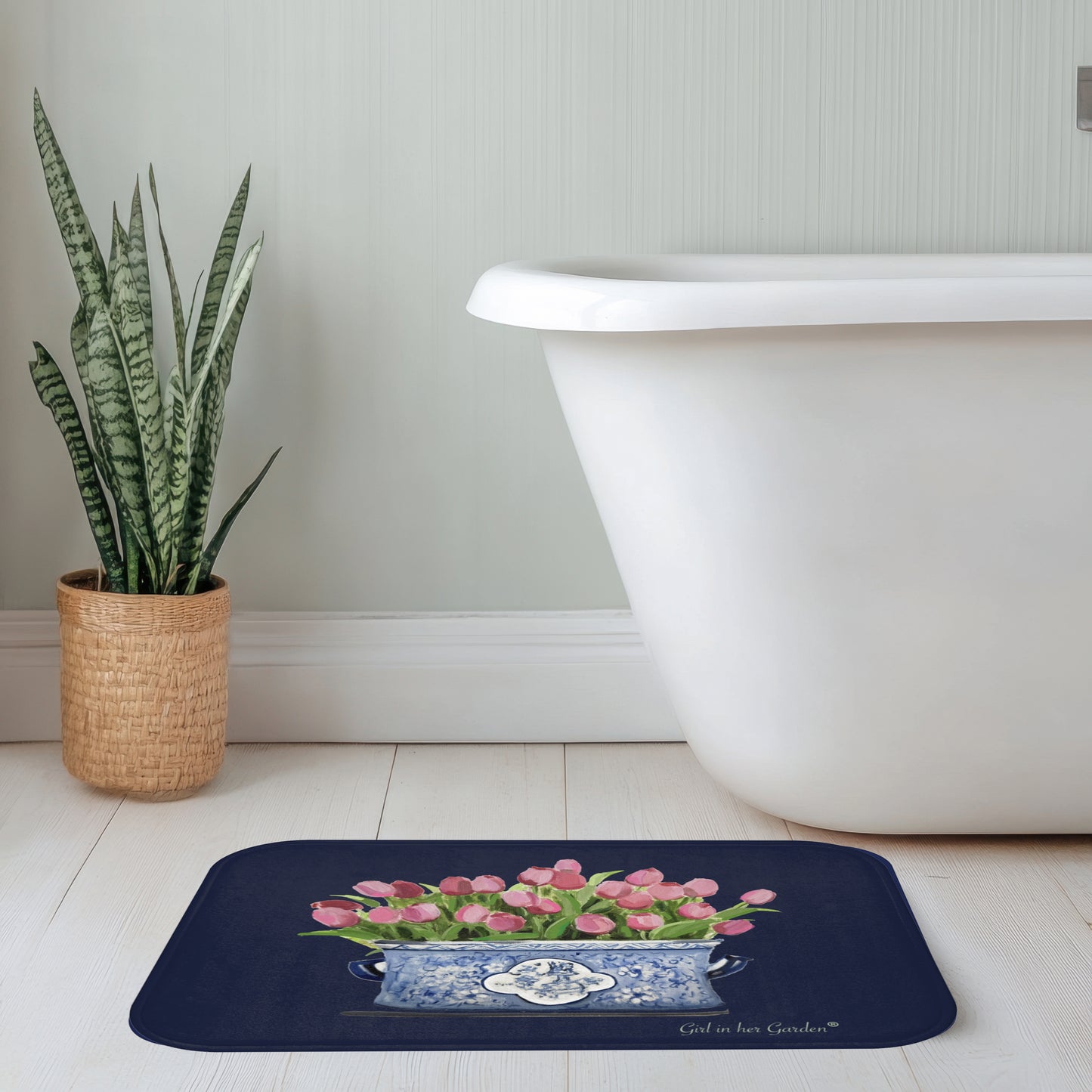 Bath mat with pink tulips design in front of a white bathtub and plant.