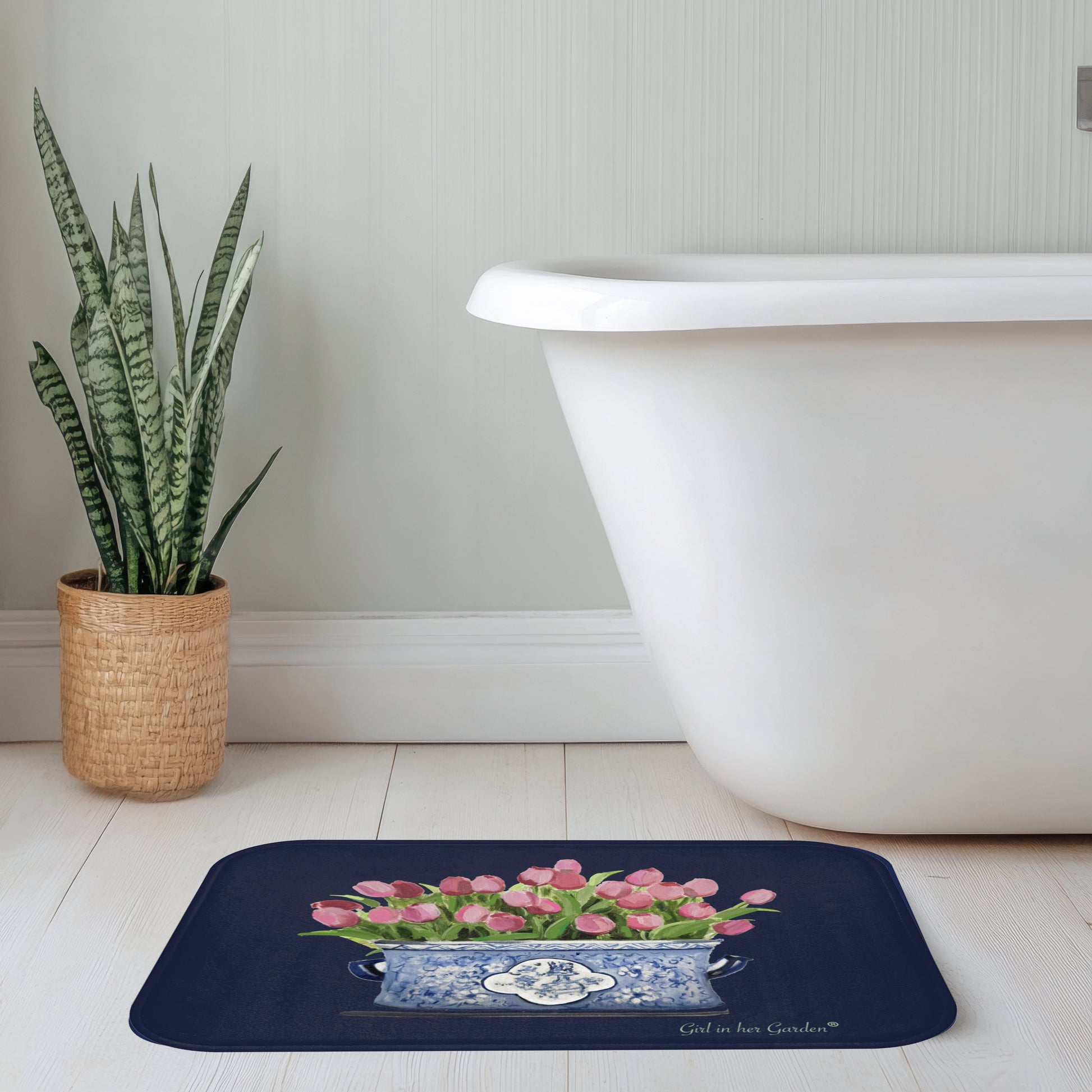 Bath mat with pink tulips design in front of a white bathtub and plant.