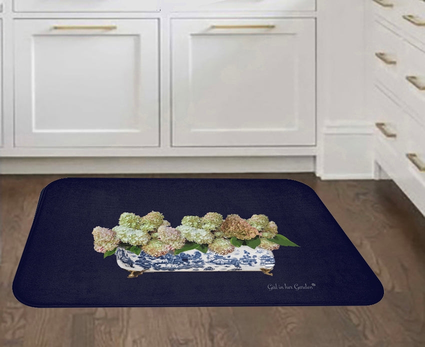 A dark navy blue rectangular bath mat with rounded corners, featuring a printed design of white and blue floral hydrangeas in a porcelain tub. The mat is shown on a brown wood floor in front of white kitchen cabinets.